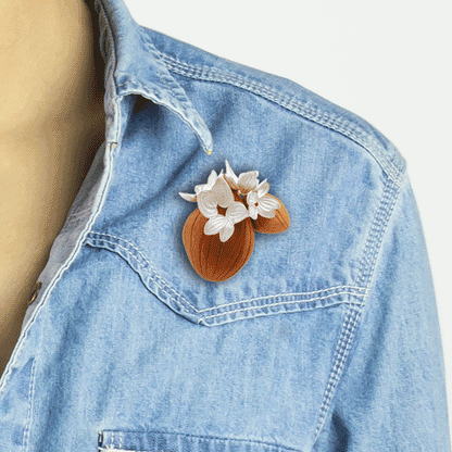 A close-up image of the 'All the Same' brooch by Erin Christensen, crafted from carved red oak with intricate sterling silver flowers adorning its surface. The silver flowers are delicately chased and repoussed, adding depth and texture to the wooden base.