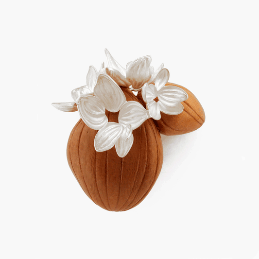 A close-up image of the 'All the Same' brooch by Erin Christensen, crafted from carved red oak with intricate sterling silver flowers adorning its surface. The silver flowers are delicately chased and repoussed, adding depth and texture to the wooden base.