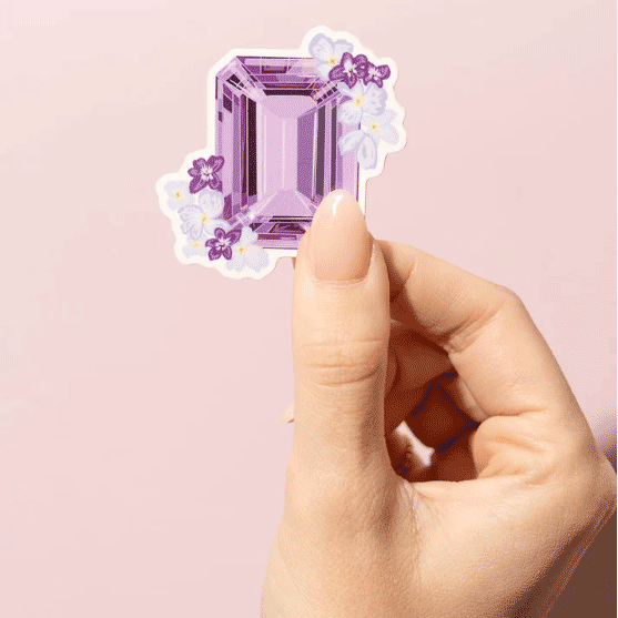 Thumb and forefinger holding a Sticker showcasing an illustrated February birthstone of amethyst and birth flowers of violets and primrose