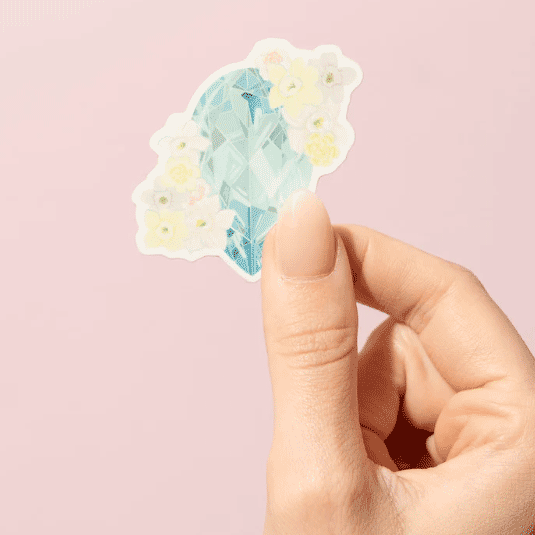Thumb and forefinger holding a Sticker showcasing an illustrated March birthstone of aquamarine and birth flowers of daffodil and jonquil.