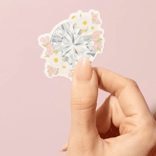 thumb and forefinger holding a 2" sticker of a diamond surrounded by daisies and sweat pea.