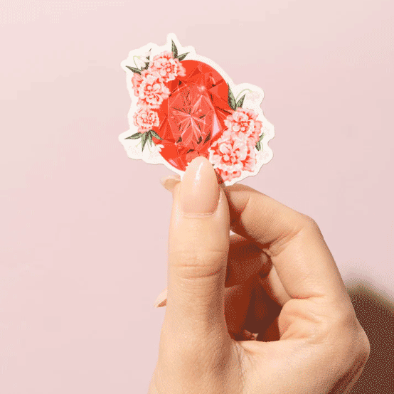 Thumb and forefinger holding a Sticker showcasing an illustrated January birthstone garnet and birth flowers of carnation and snowdrop.