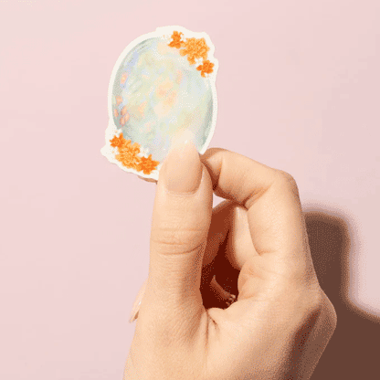 Thumb and forefinger holding a Sticker showcasing an illustrated October birthstone of opal and birth flowers of marigold and cosmos.