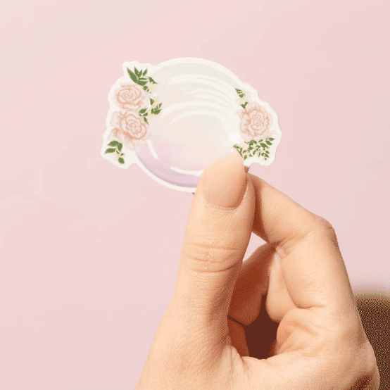 Thumb and forefinger holding a Sticker showcasing an illustrated June birthstone of pearl and birth flowers of rose and honeysuckle.