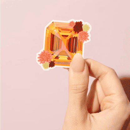 Thumb and forefinger holding a Sticker showcasing an illustrated November birthstone of topaz and birth flowers of chrysanthemum.
