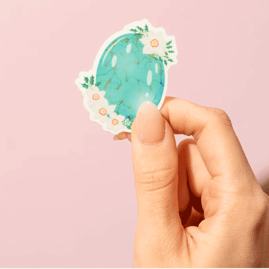 Thumb and forefinger holding a Sticker showcasing an illustrated December birthstone of turquoise and birth flowers of holly and narcissus.