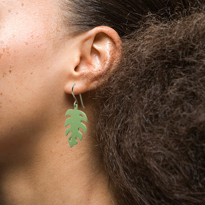 Pair of pale green drop earrings with a unique monstera leaf silhouette. The earrings are made of a glossrial and hang delicately from silver hooks. The leaf-like shape features curved edy, lightweight mateges and a pointed tip, creating a natural and organic aesthetic. These earrings are perfect for adding a touch of tropical flair to any outfit.