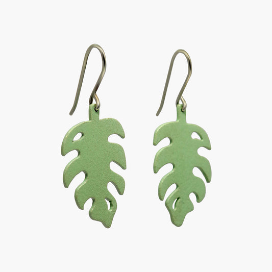 Pair of pale green drop earrings with a unique monstera leaf silhouette. The earrings are made of a glossy, lightweight material and hang delicately from silver hooks. The leaf-like shape features curved edges and a pointed tip, creating a natural and organic aesthetic. These earrings are perfect for adding a touch of tropical flair to any outfit.