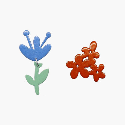 Pair of mix-and-match flower earrings in blue and orange, featuring a glossy finish and intricate detailing, with secure post backings and measuring approximately 1 inch in diameter.